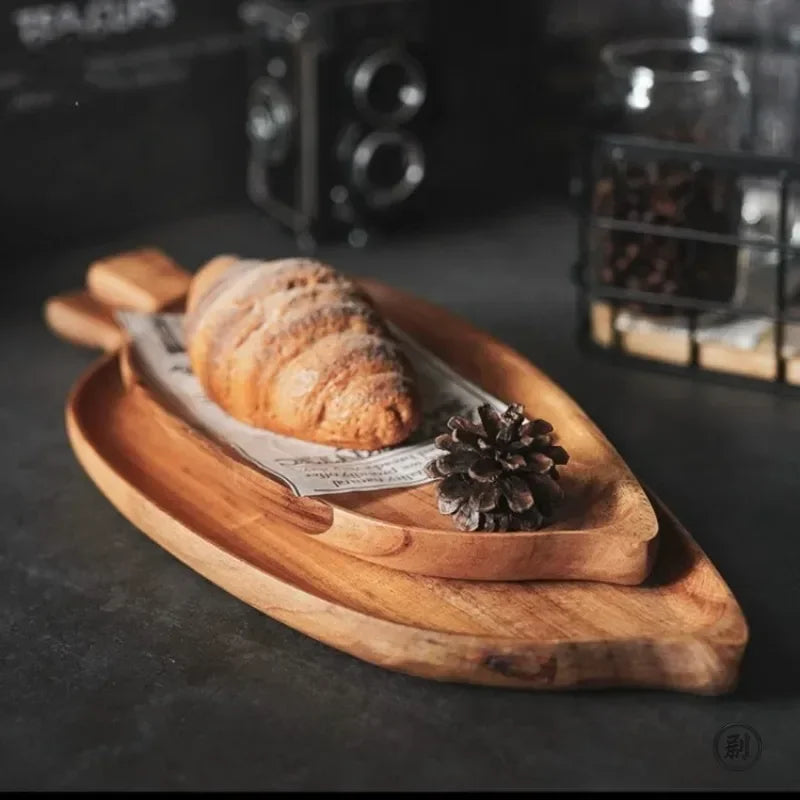 Palm leaf-shaped wooden dessert tray with croissant, pinecone decoration, and artisan details on dark surface.