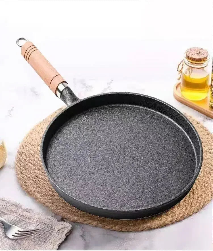 Cast iron egg skillet with wooden handle on cork mat, surrounded by olive oil bottle and kitchen linens on marble surface.