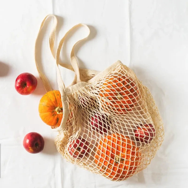 Cotton mesh grocery bag filled with apples and oranges, showing breathable net design and beige fabric handles against white 