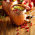 Copper Moscow mule mug with hammered finish, filled with cocktail, garnished with rosemary and pomegranate seeds on rustic wo