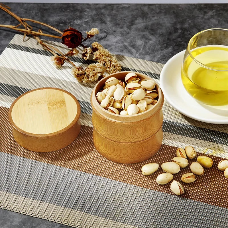 Bamboo food storage box with lid containing pistachios, displayed with olive oil, dried flowers, and scattered nuts on linen 