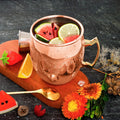 Hammered copper insulated travel mug filled with watermelon, lime, and grapefruit slices, displayed on wooden board with fres