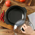 Black ceramic clay cooking pot with glass lid displayed on wooden surface, surrounded by fresh tomatoes, red chilies, and kit