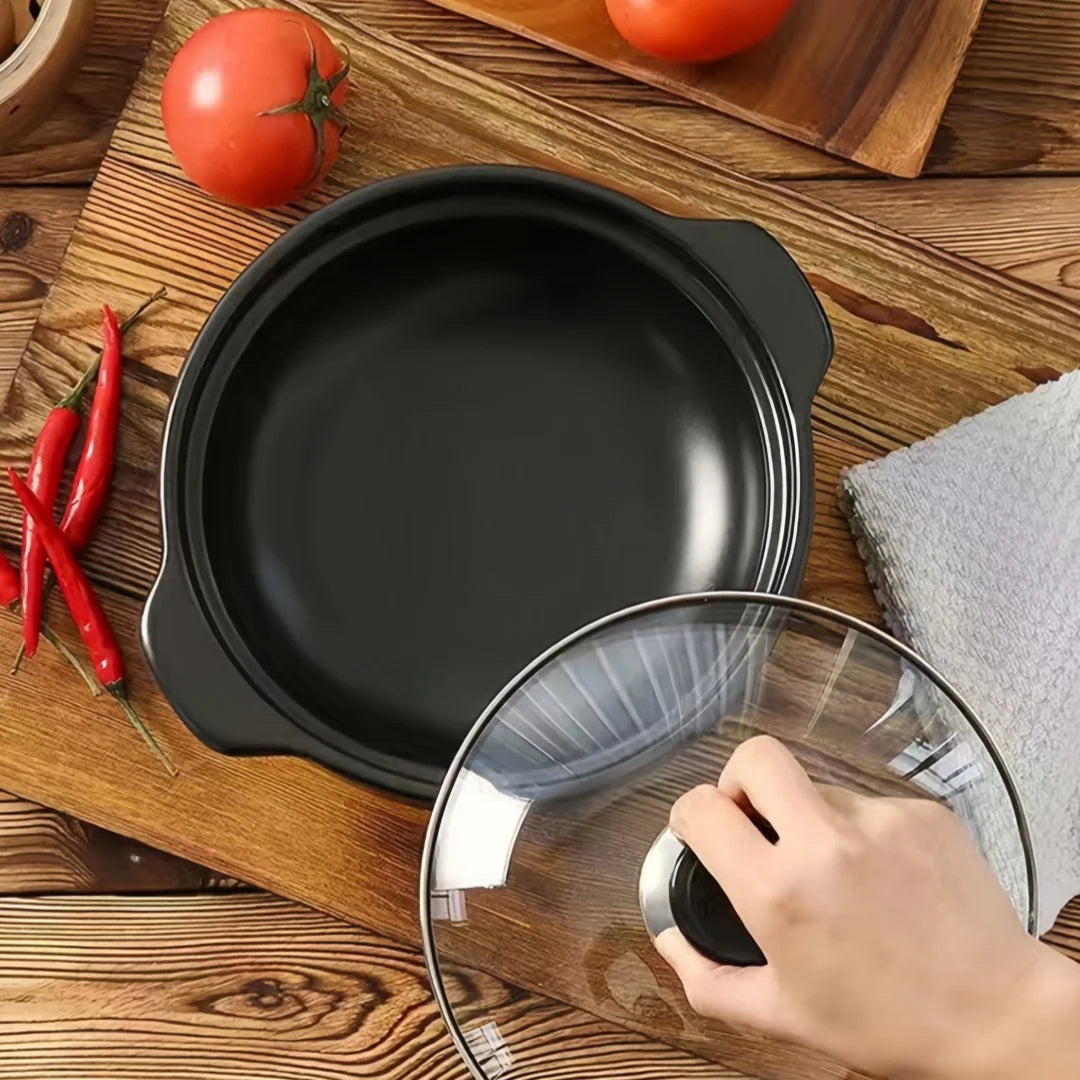Black ceramic clay cooking pot with glass lid displayed on wooden surface, surrounded by fresh tomatoes, red chilies, and kit