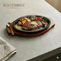Cast iron fajita griddle pan on wooden serving board with grilled steak, potatoes, and vegetables, Rootborn Rituals branding