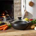 Black ceramic clay cooking pot with glass lid surrounded by fresh vegetables and herbs on rustic wooden surface, fireplace ba